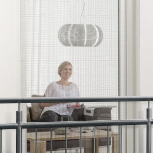 Eine Frau sitzt auf dem Sofa im Wohnraum. Sie schaut hinaus durch das Fenster am französischen Balkon, an dem der Fensterbausatz "MASTER SLIM XL" befestigt ist.