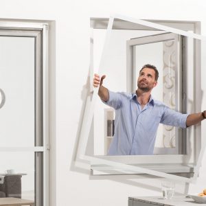 Ein Mann hält das fertig zusammengebaute Fliegengitter compact mit beiden Händen durch das geöffnete Fenster nach draußen, um es am Fensterrrahmen anzubringen.