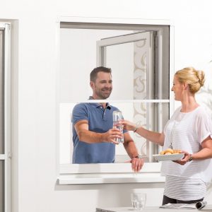 Ein Mann reicht einer Frau eine Wasserkaraffe durch das geöffnete Fenster nach draußen. Der Fensterrollo smart in weiß, der sich vor dem Fenster befindet, ist halb geöffnet.