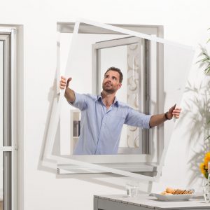 Ein Mann hält das fertig zusammengebaute Fliegengitter flächenbündig mit beiden Händen durch das geöffnete Fenster nach draußen, um es am Fensterrrahmen anzubringen.