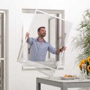 Ein Mann hält das fertig zusammengebaute Teleskopfenster mit beiden Händen durch das geöffnete Fenster nach draußen, um es am Fensterrrahmen anzubringen.